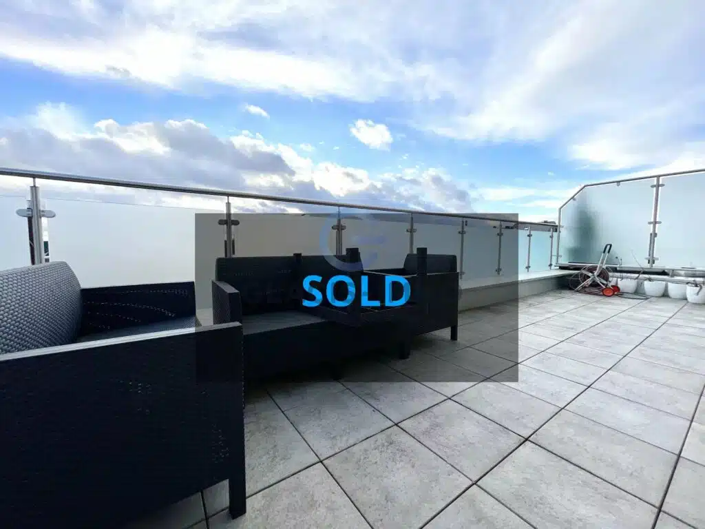 Tiled outdoor terrace furnished with dark woven seating and bordered by glass railings, with a large blue SOLD graphic across the view.